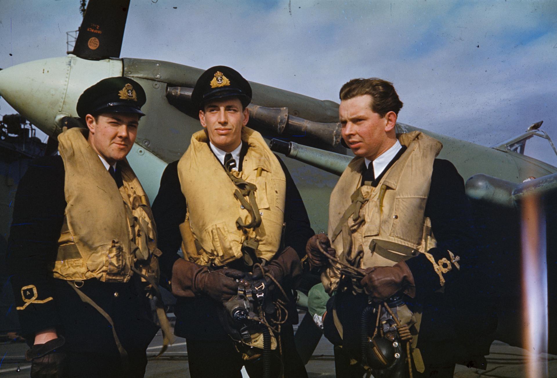 Three Fleet Air Arm pilots in yellow life jackets waiting for orders on an aircraft carrier deck, a naval fighter behind them (IWM TR 1121)